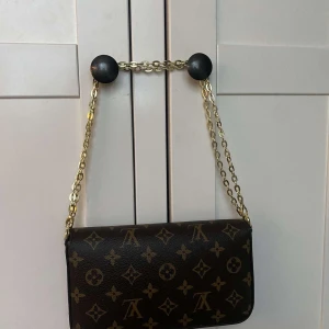 Louis Vuitton monogram axelväska - Säljer en klassisk Louis Vuitton axelväska i mörkbrunt skinn med ikoniskt monogrammönster och guldfärgad kedja. Väskan har ett stilrent kuvertformat lock med tryckknapp och är perfekt för dig som vill ha en tidlös och lyxig accessoar.