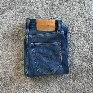 Blå jeans från Tiger of Sweden - Snygga blå jeans från Tiger of Sweden med klassisk femficksdesign och orange kontrastsömmar. Jeansen har normal midja och raka ben, tillverkade i mjukt bomullsjeansmaterial. Perfekta för en stilren och avslappnad look.