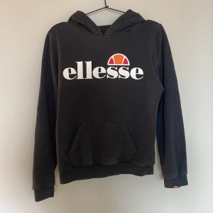 Svart Ellesse hoodie med logga storlek 152-158  - Svart hoodie från Ellesse med stor vit och orange logga på bröstet. Liten Ellesse-detalj på ärmslutet för extra stilpoäng. Strl 152-158. ( 12-13 år ) 