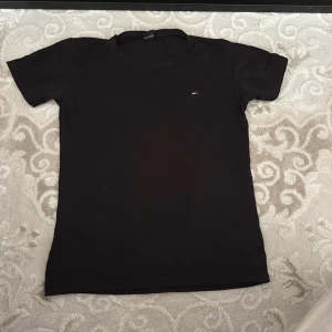 Svart t-shirt från Tommy Hilfiger - Svart t-shirt från Tommy Hilfiger med klassisk passform och liten broderad logga på bröstet. T-shirten har rund halsringning och korta ärmar, perfekt för en clean och stilren look. Det är en storlek XL men jag som är en storlek S kan ha på mig den.