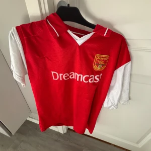 Arsenal Henry 14 fotbollströja röd/vit M/L - Klassisk Arsenal fotbollströja med Dreamcast-tryck och broderat klubbmärke. Röd med vita ärmar, kortärmad och med Henry 14 på ryggen. Perfekt för dig som älskar retrostil och vill hylla en av klubbens största legender.