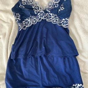 Marinblå linnetopp och shorts  med spetsdetaljer - Säljer en marinblå linnetopp och short tillhörande med vita spetsdetaljer i blommigt mönster över bysten och axelbanden. Toppen är ärmlös och har en mjuk, flowy passform. Materialet är polyester med lite stretch, perfekt för en chill och snygg look.