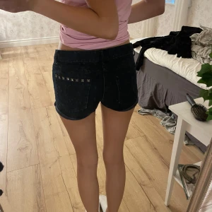 Svarta jeansshorts med nitar - Snygga svarta jeansshorts med uppvikta ben och coola silverfärgade nitar vid fickorna fram och bak. Klassisk femficksmodell med knapp och dragkedja. Perfekta för dig som gillar en edgy stil och vill ha något som sticker ut lite extra.