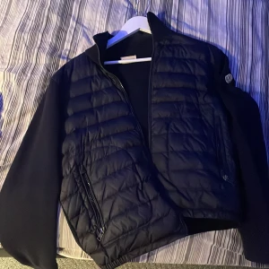 Blå Moncler cardigan med dragkedja - Snygg blå cardigan från Moncler köpt i USA 2017 med quiltad framsida och ärmar i stickad ull. Jackan har hög krage, två fickor med dragkedja och klassisk Moncler-logga på ärmen. Perfekt för dig som vill ha en stilren och exklusiv look. Priset kan ändras vid smidig affär. Kvitto finns