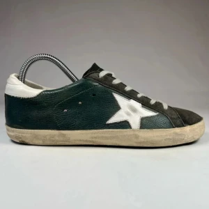 Svarta sneakers golden goose - Snygga svarta sneakers från Golden Goose med vit stjärndetalj på sidan och vit häl. Skorna har grå snörning, rund tå och slitna detaljer för en cool, vintage look. Ovandelen är i skinn och mocka, perfekt för dig som gillar streetstyle.