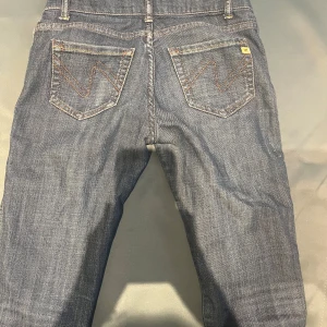 Blå jeans Wrangler med slitna detaljer - Säljer ett par klassiska blå jeans från Wrangler med raka ben och snygga slitningar nertill. Jeansen har ikoniska bakfickor med W-söm och gult märke. Tillverkade i robust denim som ger en cool vintagekänsla.