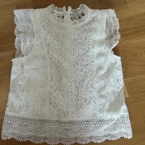 Vit spetsblus - Superfin vit blus från ONLY i spets med blommigt mönster och volangdetaljer längs ärmhålen. Hög hals med dragkedja bak och vackra broderade kanter. Perfekt för dig som gillar romantisk och detaljerad stil.