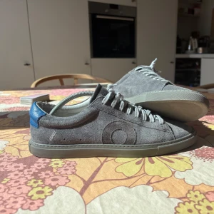 Grå Oliver cabell skor med blå detalj - Snygga sneakers i grå mocka med diskret cirkeldetalj på sidan och blå häl. Skorna har grå snörning och platt sula i matchande grått. Perfekta för dig som gillar stilrena och moderna skor med en twist. Storlek 42 knappt använda 