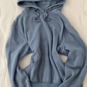 Blå hoodie från H&M Basic - Mysig ljusblå hoodie från H&M Basic med huva och snörning. Tröjan har en klassisk känguruficka framtill och ribbade muddar vid ärmslut och nederkant. Perfekt för chill dagar eller när du vill ha en enkel och stilren look.