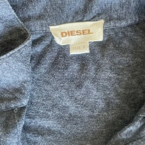 Grå långärmad tröja från Diesel S - Mörkgrå långärmad tröja från Diesel i mjuk bomullsmix. Tröjan har en unik vit printdetalj nertill, v-ringad hals med två knappar och avslappnad passform. Perfekt för dig som gillar streetstyle och vill ha något lite annorlunda.