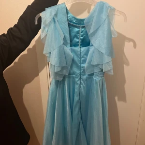 Ljusblå långklänning med volanger - Superfin ljusblå långklänning med flera lager volanger upptill och glittrande detaljer i midjan. Klänningen har ärmlös design och ett flowy tyg som ger en drömmig känsla. Perfekt för dig som vill sticka ut med en sagolik look.