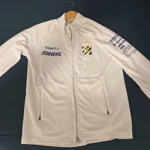 Vit träningsjacka IFK Göteborg Craft - Vit långärmad träningsjacka från Craft med IFK Göteborgs klubbmärke på bröstet och flera sponsorloggor. Jackan har dragkedja framtill och två sidofickor med dragkedja. Perfekt för dig som gillar fotboll och vill representera laget.