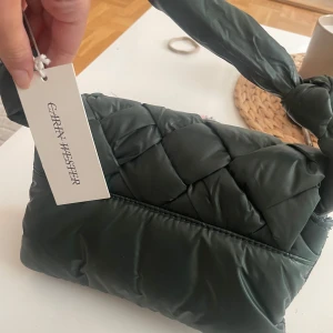  grön axelväska från carin wester - Snygg quiltad axelväska från GANNI i mörkgrönt syntetmaterial. Väskan har en vadderad, knutbar axelrem och magnetlås. Perfekt storlek för det viktigaste och ger en cool vibe till din outfit. Helt oanvänd med prislapp kvar