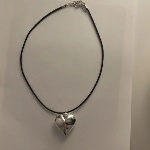 Halsband med silverfärgat hjärta - Stilrent halsband med svart snöre och ett stort, blankt hjärtformat hänge i silverfärg. Justerbart spänne i metall. Perfekt för dig som gillar minimalistiska smycken med en touch av romantik.