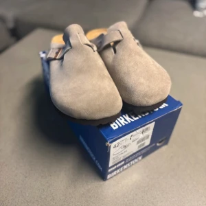 Birkenstock Boston grå mockasandaler - Grå Birkenstock Boston sandaler i mjuk mocka med justerbart spänne i metall och klassisk korksula. Skorna har en stängd tå och öppen häl, perfekt för chill vardagsstil. Ikonisk komfort och snygg minimalistisk look.