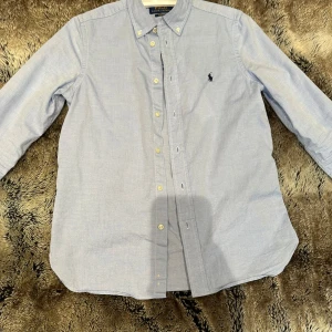 Ljusblå skjorta Polo Ralph Lauren - Klassisk ljusblå skjorta från Polo Ralph Lauren i slim fit-modell. Skjortan har button down-krage, långa ärmar och broderad logga på bröstet. Tillverkad i mjuk bomull och stängs med knappar framtill. Perfekt för en clean och stilren look. Sitter lite mindre i storleken