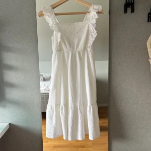 Vit långklänning med volang från Zara - Superfin vit långklänning från Zara med broderade detaljer och volang vid axelbanden. Klänningen har en luftig passform, smock i ryggen och är tillverkad i ett lätt bomullstyg. Perfekt för sommarens alla tillfällen!