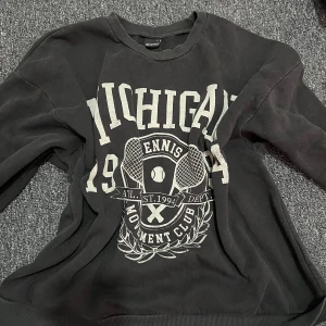 Svart sweatshirt från Gina Tricot - Cool svart sweatshirt från Gina Tricot med stort Michigan-tryck och tennismotiv framtill. Tröjan har rund hals, långa ärmar och ribbade muddar. Perfekt för en sportig och avslappnad look.