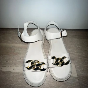 Chunky vita sandaler med guldkedja - Snygga chunky sandaler i vitt med breda remmar och en stor guldfärgad kedjedetalj framtill. Sandalerna har grov sula och justerbart spänne vid ankeln. Perfekta för att lyfta din sommarstil med en trendig touch.