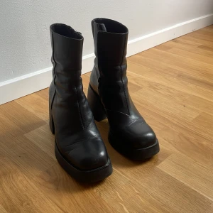 Svarta boots med klack från Vagabond - Snygga svarta boots från Vagabond i skinn med chunky klack och platt sula. Modellen har rund tå, dragkedja på insidan och en stilren, minimalistisk look. Perfekta för dig som gillar en edgy vibe och vill ha något som sticker ut.