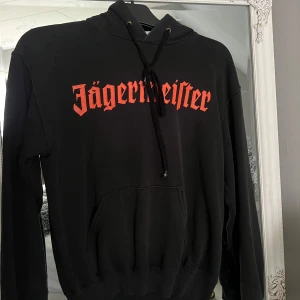 Svart Jägermeister hoodie med tryck - Svart hoodie från Jägermeister med stort orange tryck framtill och bak, där bakstycket har det klassiska hjort- och korsmotivet. Tröjan har huva med snörning och en stor magficka. Perfekt för dig som gillar streetwear och vill sticka ut.