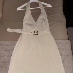 Beige skjort klänning med bälte - Supersnygg beige skjort klänning med halterneck och markerad midja tack vare ett brett bälte med guldspänne. Klänningen har djup v-ringning och är ärmlös, perfekt för varma dagar. Materialet är lätt och har en diskret struktur.