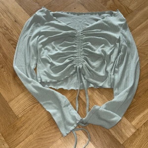 Ljusgrön croppad ribbad topp - Trendig ljusgrön croppad topp med långa ärmar och snygg snörning framtill som ger en rynkad effekt. Toppen har v-ringning och är gjord i ett ribbat material som känns mjukt mot huden. Perfekt för dig som gillar en stilren men ändå cool look.