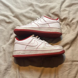 Nike Air Force 1 vit/röd sneakers - Nike Air Force 1 sneakers i vitt skinn med röda detaljer och röd sula. Klassisk låg modell med snörning, perforerad tå och röd Nike-logga på häl och plös. Insidan är fodrad i rött tyg och skon har en platt sula för skön streetstil.