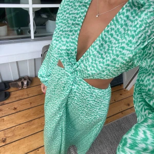 Grön och vit långklänning med cut-outs - Säljer en snygg långklänning i grönt och vitt mönster med djup v-ringning, knutdetalj framtill och cut-outs i midjan. 
