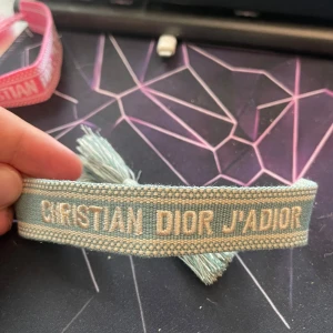 Blått Christian Dior J'Adior armband - Supersnyggt blått armband från Christian Dior med texten 'J'Adior' broderad i vitt. Armbandet har fransar i ändarna och är gjort i ett mjukt tygmaterial. Perfekt accessoar för att lyfta din outfit.