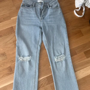 Ljusblå raka jeans med slitningar - Säljer ett par ljusblå jeans med raka ben och snygga slitningar över knäna. Klassisk femficksmodell med hög midja och dragkedja. Jeansen är i ett mjukt bomullsmaterial och har en avslappnad, trendig vibe.