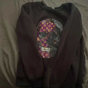 Svart sweatshirt med lv skalle tryck  - Cool svart sweatshirt med ett stort tryck av en döskalle framtill, dekorerad med lila och orange mönster. Tröjan har rund hals och långa ärmar, perfekt för dig som vill sticka ut med en edgy stil Louis vuitton.
