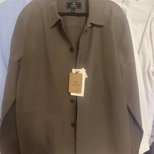 Brun overshirt från Matinique - Stilren brun overshirt från Matinique med klassisk krage, knäppning framtill och två stora fickor. Perfekt lager-på-lager-plagg med enkel design och skön passform. Tillverkad i mjuk bomull för en avslappnad vibe. Helt oanvänd! Nypris ca 1500