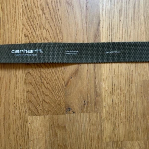 Grönt Carhartt bälte i polyester - Snyggt grönt bälte från Carhartt med klassisk metallspänne och logga på spännet. Bältet är gjort i slitstark polyester och har en enkel, stilren design som passar till många outfits. One size och lätt att justera längden.