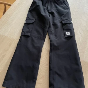 Svarta skidbyxor från WearColour - Svarta skidbyxor från WearColour med bred passform och stora fickor på sidorna. Byxorna har knapp- och dragkedjestängning samt diskret logga på ena benet. Möjlughet att reglera midjebredden med kardborre.
