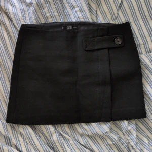 Svart kortkjol från MNG med knappdetalj - Stilren svart kortkjol från MNG i italienskt tyg. Kjolen har en rak passform och snygg knappdetalj framtill samt dold dragkedja i sidan. Perfekt att matcha med en cool topp eller oversized jacka för en trendig look.