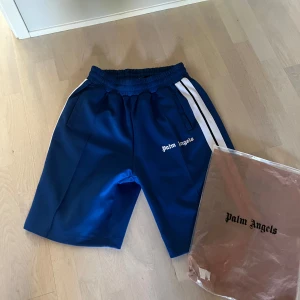 Blå Palm Angels shorts - Snygga blå shorts från Palm Angels med vita ränder längs sidorna och broderad logga framtill. Modellen har elastisk midja , perfekta för dig som gillar streetwear. 