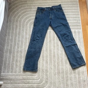 Levi's 501 blå jeans W31 L32 - Klassiska blå Levi's 501 jeans med rak passform och knappgylf. Jeansen har fem fickor, snygga kontrastsömmar och den ikoniska läderpatchen bak i midjan. Perfekta för dig som gillar tidlös denimstil.