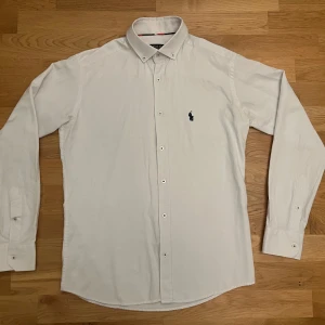 Vit skjorta från Polo Ralph Lauren - Klassisk vit skjorta från Polo Ralph Lauren med button down-krage och broderad marinblå logga på bröstet. Skjortan har lång ärm, knappar framtill och är tillverkad i bomull. Perfekt för en clean och stilren look.