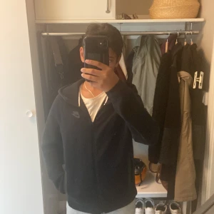Svart Nike hoodiejacka med dragkedja - Svart hoodiejacka från Nike med hel dragkedja framtill och två fickor med dragkedja. Jackan har huva och diskret Nike-logga på bröstet. Tillverkad i mjukt bomullsmaterial, perfekt för chill dagar eller till träning. Knappt använd! Så väldigt bra skick😁👍Priset är inte hugget i sten!