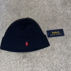 Mörkblå stickad mössa Polo Ralph Lauren - Snygg mörkblå stickad mössa från Polo Ralph Lauren med klassisk röd broderad logga framtill. Mössan har bred uppvikt kant och är tillverkad i mjukt material som håller dig varm och stilren hela vintern.