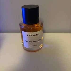 ESSNCE Orange Blossom Eau de Parfum - ESSNCE Orange Blossom är en blommig gourmanddoft med toppnoter av bergamott, neroli och rosépeppar. Hjärtat har kaprifol, ros, apelsinblomma, iris och jasmin. Basen är söt med socker, vanilj, karamell, amber och mysk. Flaskan är rund i glas med svart lock och bär en stilren etikett.