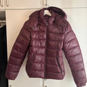 Vinröd pufferjacka lager 157 - Säljer en vinröd pufferjacka från Nikki i storlek S. Jackan har huva, dragkedja framtill och två fickor med dragkedja. Den är quiltad och har en snygg, lite glansig finish. Perfekt för kalla dagar när du vill hålla stilen.