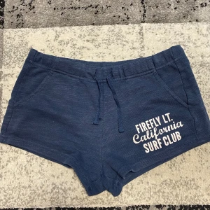 Blå shorts från Firefly - Mörkblå shorts från Firefly med trycket 'California Surf Club' på ena benet. Mjukt bomullsmaterial, elastisk midja med snörning och två fickor fram. Perfekta för sommardagar eller chilla hemma.