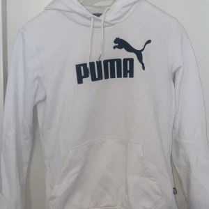 Vit hoodie från Puma med logga - Vit hoodie från Puma med stor svart logga och puma-tryck på bröstet. Klassisk känguruficka framtill, snörning i huvan och mjukt material. Perfekt för en chill och sportig look. 