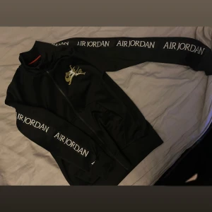 Svart Air Jordan jacka med dragkedja - Svart jacka från Nike Air Jordan med vit logga på bröstet och texten 'AIR JORDAN' längs båda ärmarna. Jackan har hel dragkedja framtill, hög krage och två fickor. Perfekt för dig som gillar sportig och clean stil.