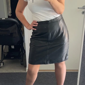 Svart skinnkjol i klassisk modell - Säljer en svart skinnkjol med rak passform och midjehög linning. Kjolen är knälång.Perfekt för dig som vill ha en edgy och tidlös look. Passar lika bra med boots som sneakers.Använd bara vid ett tillfälle några timmar Läder Storlek 42, måttet  Höften 102 Midja 80 Längd 52