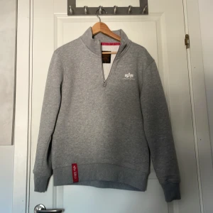 Grå half zip hoodie Alpha Industries - Snygg grå hoodie från Alpha Industries med half zip och broderad logga på bröstet. Mjuk insida i fleece och klassisk passform. Långärmad modell med ribbade muddar och röd detalj nertill. Perfekt för kyliga dagar. Pris kan diskuteras⚜️⚜️