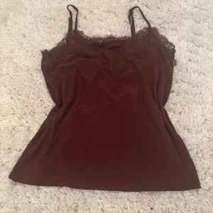 Vinröd linnetopp med spetsdetaljer - Säljer en vinröd linnetopp med tunna axelband och vacker spets längs urringningen. Toppen har en mjuk, slät yta och en lätt glans.