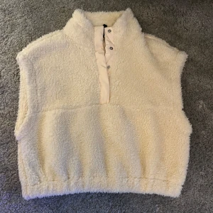 Beige teddyväst från H&M Divided - Mysig beige teddyväst från H&M Divided i storlek S. Västen har hög krage, tryckknappar framtill och är tillverkad i fluffigt fleecematerial. Perfekt att slänga över en hoodie eller långärmad tröja för extra värme och stil.